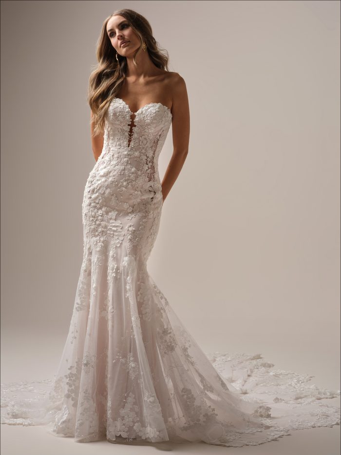 Bride in a romantic strapless fit-and-flare wedding dress adorned with floral lace motifs, illusion side panels, and a dramatic scalloped train, posing with hands in pockets.