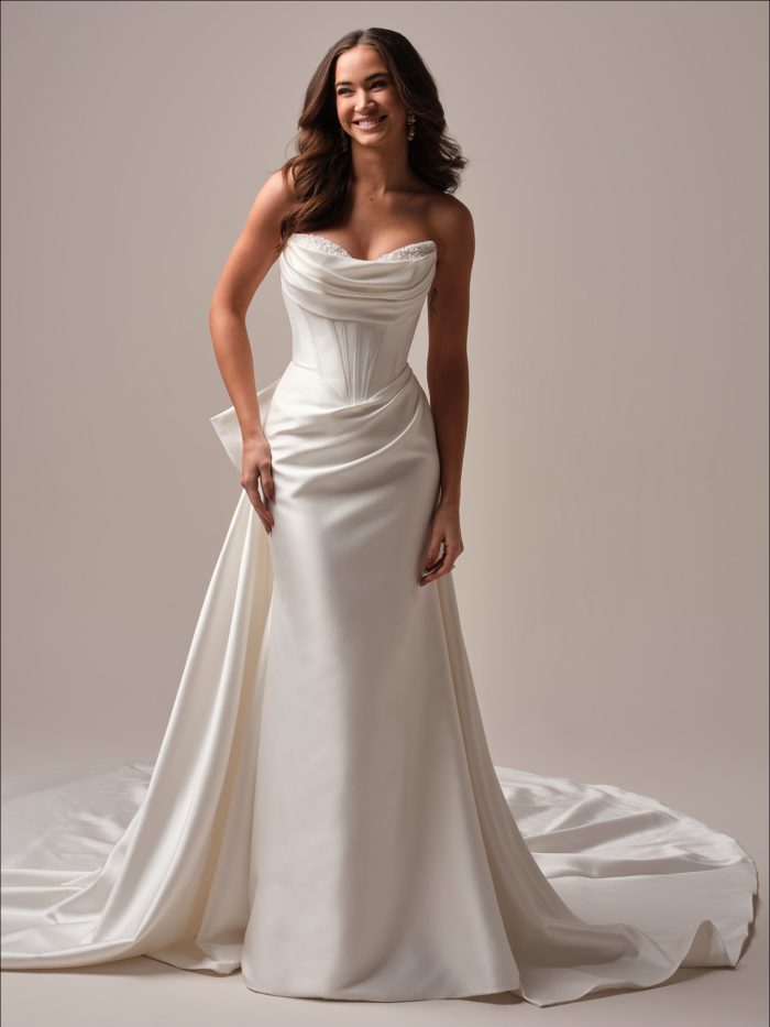 Front view of a strapless fit-and-flare wedding dress with a draped satin neckline, corset-style bodice, and side-draped skirt flowing into a chapel-length train.