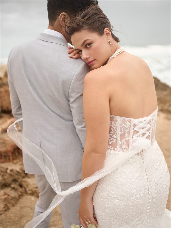 Back view of a plus size bride in a strapless lace fit-and-flare gown with a lace-up corset and button details, leaning on her groom in a light gray suit by the ocean.