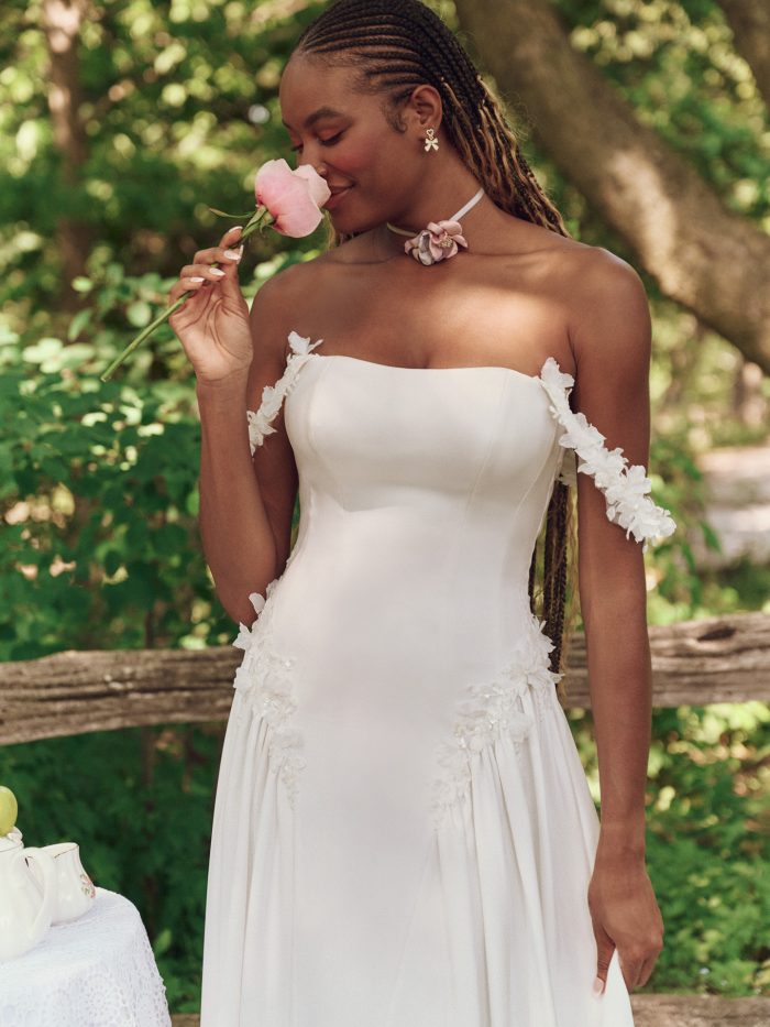 Close-up of a bride in the Maggie Sottero Morwenna gown, gently smelling a pink rose. The image highlights the floral off-the-shoulder straps, clean neckline, and delicate 3D embellishments along the bodice in a dreamy outdoor setting.