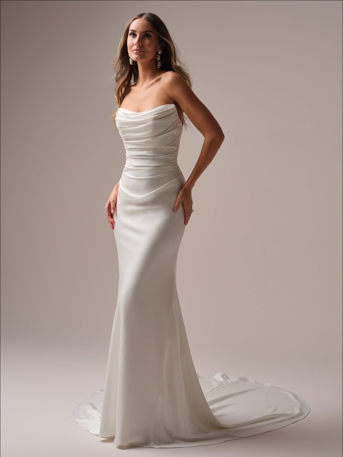 Full-length view of a bride wearing a strapless satin sheath wedding dress with a softly draped bodice, figure-hugging silhouette, and sweeping train with button detail.