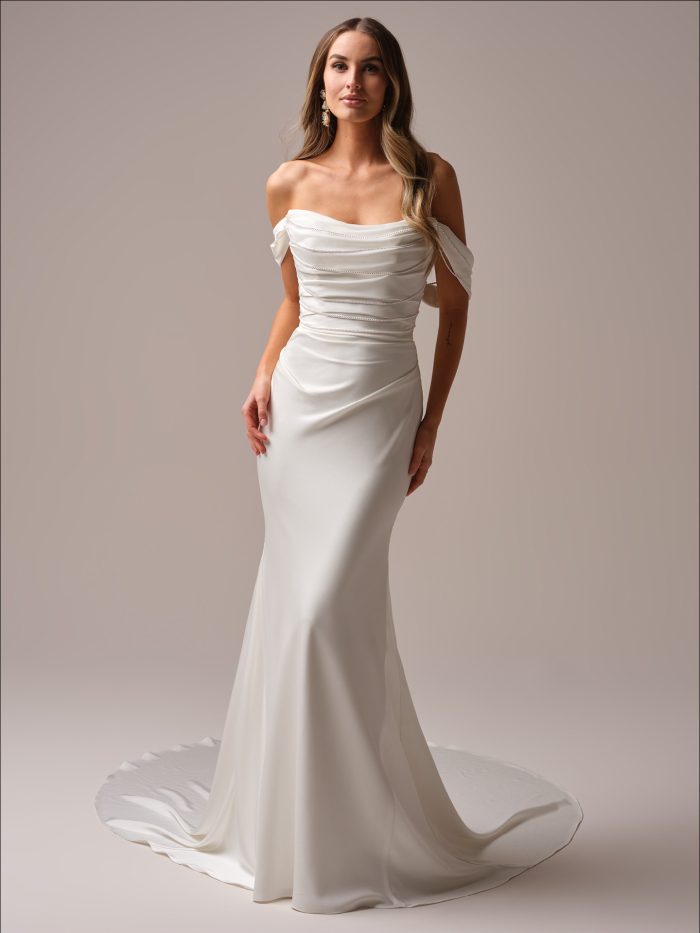 A bride wears the Maggie Sottero Rhoda sheath wedding dress, featuring a draped off-the-shoulder neckline with pearl detailing, a fitted satin silhouette, and a minimalist train for a sleek, modern look.