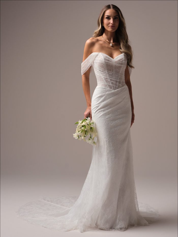 Full-length view of a bride in an off-the-shoulder sheath wedding dress featuring a ruched lace bodice, illusion sleeves, and a soft chapel-length train.