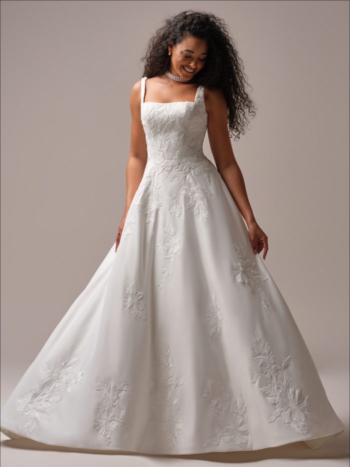 A bride wearing the Maggie Sottero Sheridan A-line wedding dress, featuring a square neckline, wide straps, and botanical embroidery on Mikado fabric. The full A-line skirt flows elegantly to the floor.