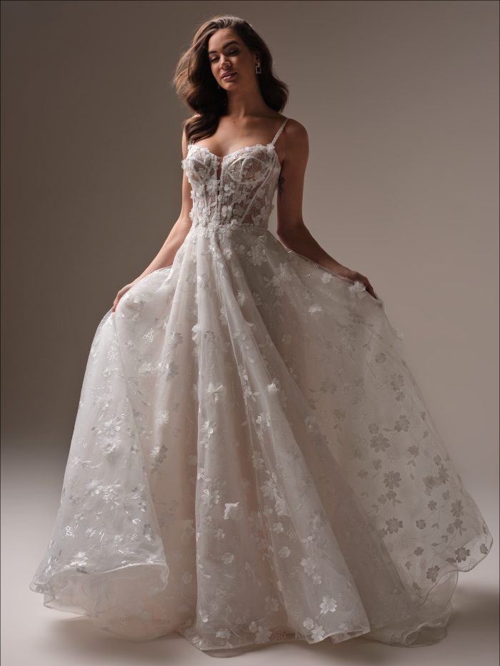 Bride in a floral lace ballgown wedding dress with a sheer corset bodice, delicate spaghetti straps, and button-back detail, holding the side of her gown.