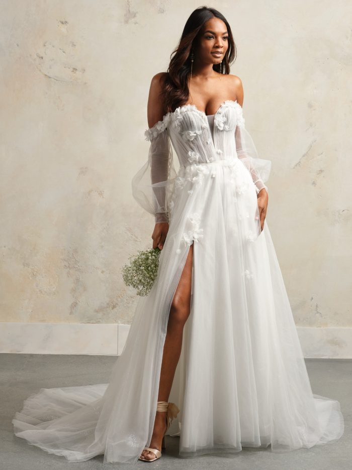 Bride wearing a romantic off-the-shoulder A-line wedding dress with sheer corset bodice, 3D floral appliqués, long illusion sleeves, and a high slit skirt with a flowing train.