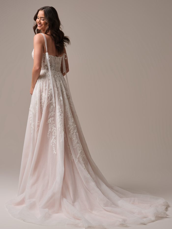 Back view of an A-line wedding dress with delicate lace embroidery, soft blush tulle layers, and dainty ribbon tie straps flowing from the shoulders