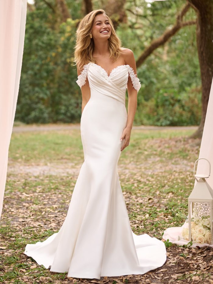 A bride wears the Rebecca Ingram Cindy fit-and-flare wedding dress, featuring a sweetheart neckline with off-the-shoulder floral lace straps and ruched bodice, standing in an outdoor garden setting framed by blush drapery.