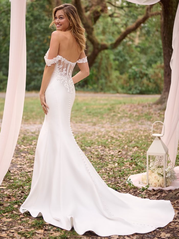 Back view of the Rebecca Ingram Cindy gown showcasing its illusion lace bodice, floral off-the-shoulder straps, fabric-covered buttons, and a softly flared train in a romantic outdoor ceremony setup.