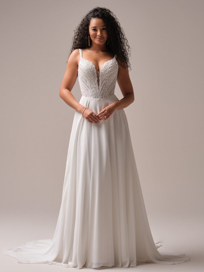 A bride wears the Rebecca Ingram Clara A-line wedding dress, featuring a plunging V-neckline, beaded lace bodice, and soft chiffon skirt, creating a romantic and flowy bridal look.