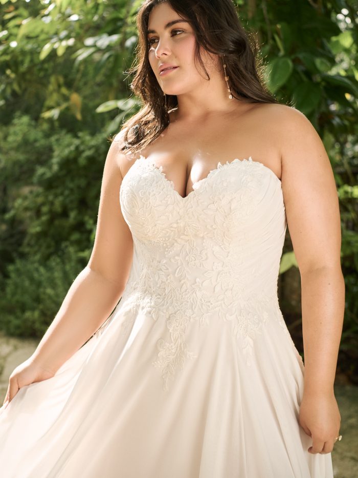 Plus-size bride in a strapless chiffon A-line gown with a lace bodice and thigh-high slit, walking across a rustic stone pathway toward a wooden gazebo.