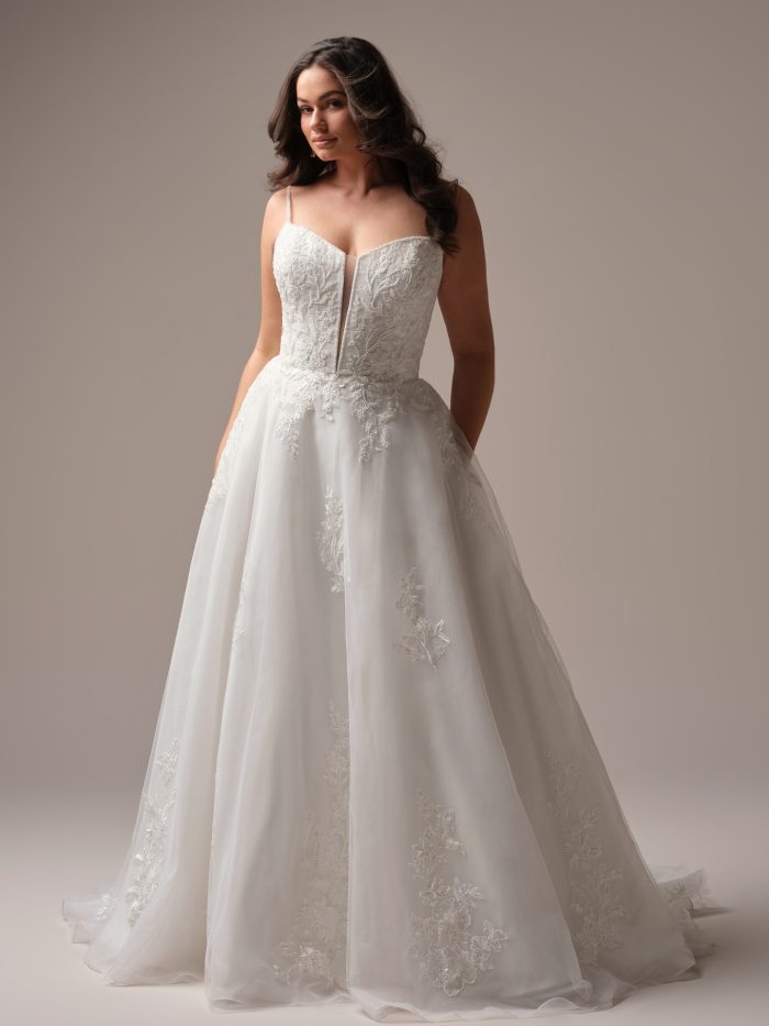 Bride wearing a ballgown wedding dress with beaded lace bodice, plunging illusion neckline, and flowing tulle skirt, posing with hands in pockets.