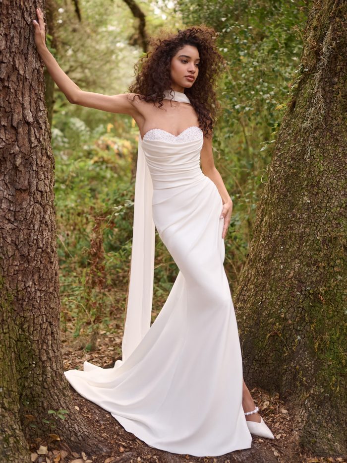 A bride wearing the Rebecca Ingram Kathleen sheath wedding dress poses between two trees in a forest, showcasing the gown’s elegant draping, pearl-embellished sweetheart neckline, and flowing scarf detail.