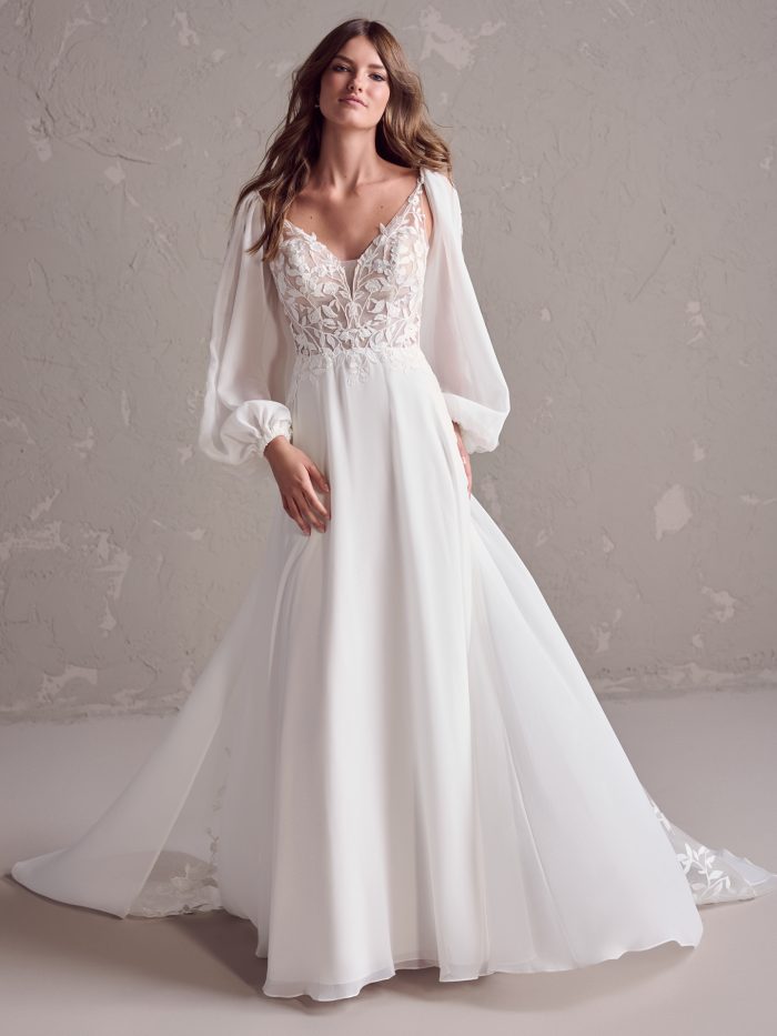 Bride wearing a sheath wedding dress with a lace bodice, plunging neckline, and long billowing chiffon sleeves, paired with a flowing skirt and train.