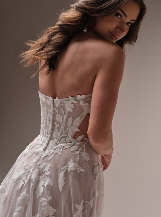 Back view of a floral A-line wedding dress featuring a strapless illusion corset bodice, fabric-covered buttons, and dimensional floral appliqués trailing down a soft tulle skirt.