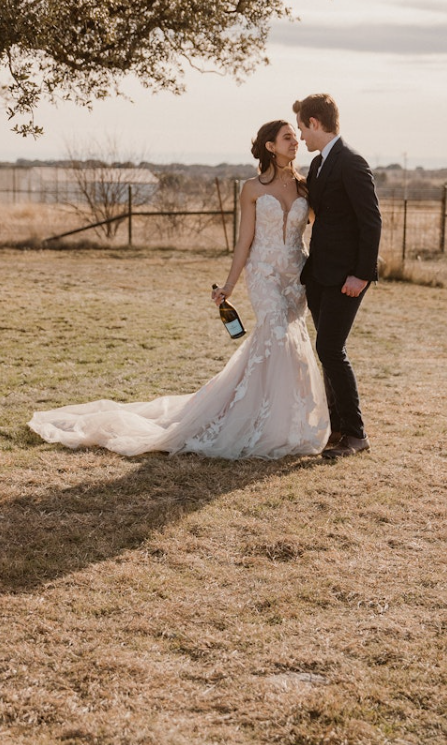 Bride wearing a boho bridal dress with a strapless sweetheart neckline, lace fit-and-flare silhouette, and flowing train, holding a champagne bottle while standing outdoors with the groom in a dark suit.