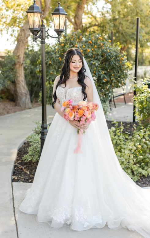 Bride in a lace A-line wedding gown with a sweetheart neckline and long train, holding a bright bouquet of pink and orange flowers, posing outdoors under a vintage lamp post.