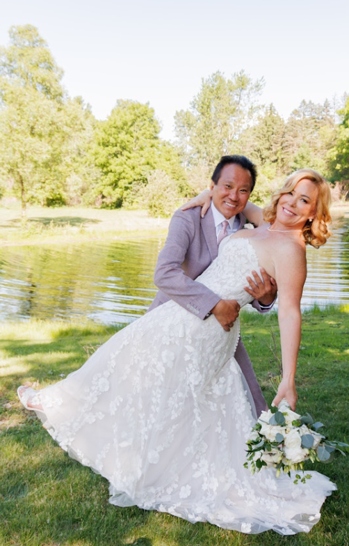 Bride in a strapless lace boho bridal dress with a flowing A-line skirt, holding a bouquet of white roses while playfully leaning back in the groom’s arms near a lakeside.