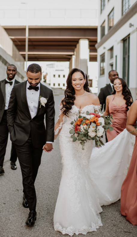 Bride in an off-the-shoulder lace wedding gown with long illusion sleeves and a fitted silhouette, carrying a vibrant bouquet while walking hand-in-hand with the groom in a black tuxedo, surrounded by their wedding party.
