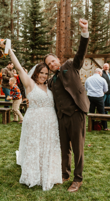 Bride in a lace A-line boho bridal dress with a V-neckline and floral appliqués, celebrating outdoors with the groom in a brown suit at a rustic forest wedding.