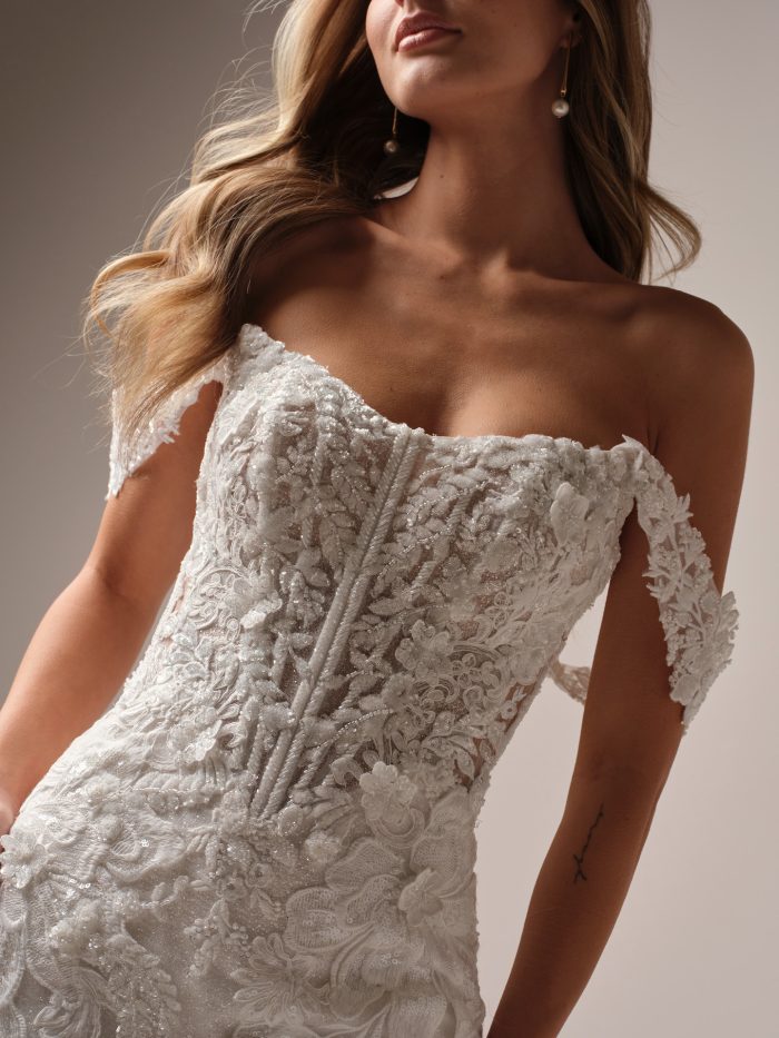 Close-up of a fit-and-flare wedding dress featuring off-the-shoulder straps, illusion lace bodice with exposed boning, and intricate beaded floral embroidery.