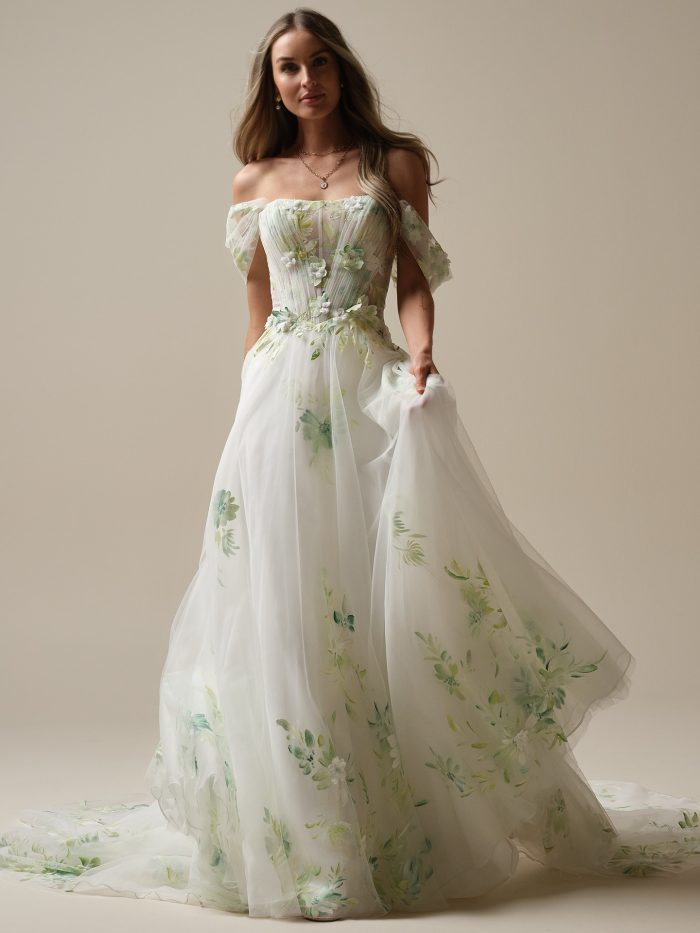 Bride wearing a romantic off-the-shoulder A-line wedding dress with a sheer corset bodice and flowing skirt adorned with watercolor-style green floral designs.