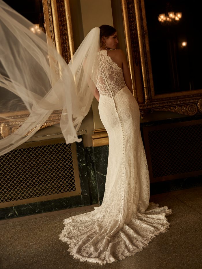 Back view of a bride wearing the Anya sheath wedding dress by Maggie Sottero, showcasing the lace halter bodice, button-back detail, and flowing lace train, standing in an ornate gilded interior.