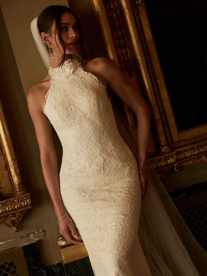 Close-up of a bride wearing the Anya sheath wedding dress by Maggie Sottero, featuring a lace halter neckline with floral detail, standing in an ornate gilded interior.