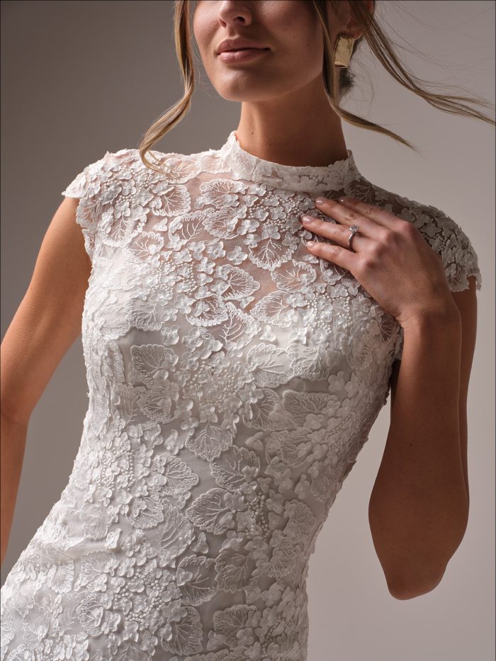 Close-up of the Maggie Sottero Auburn sheath wedding dress showcasing the illusion lace high neckline, delicate cap sleeves, and intricate floral lace motifs with subtle 3D texture.