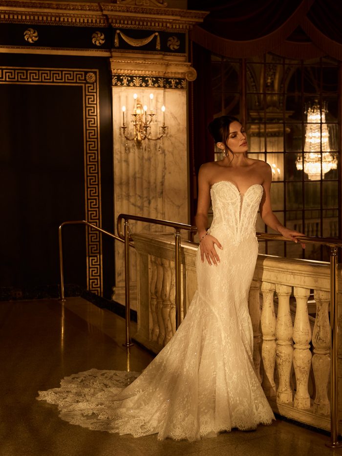 Bride wearing the Bellevue Royale fit-and-flare wedding dress by Maggie Sottero, featuring a beaded plunging sweetheart bodice and sparkling lace trumpet skirt, standing on a balcony in an ornate theater.