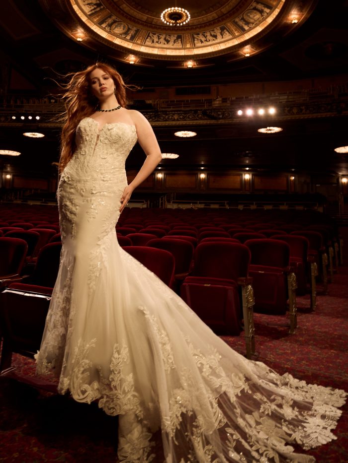 Bride wearing the Estelle fit-and-flare wedding dress by Maggie Sottero, featuring a strapless floral lace bodice and dramatic lace-trimmed train, standing in an ornate theater.