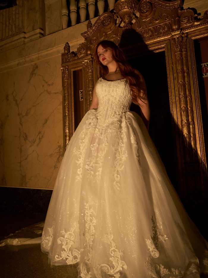 Bride wearing the Lyra ballgown wedding dress by Maggie Sottero, featuring a lace-appliquéd sparkle tulle skirt and fitted bodice, standing beside an ornate gilded doorway.