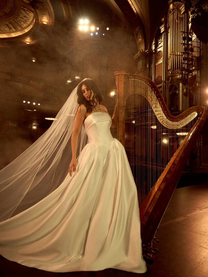 Bride wearing the Rane ballgown wedding dress by Maggie Sottero, featuring a structured strapless bodice and full satin skirt with flowing cathedral veil, standing beside a harp in an ornate theater.