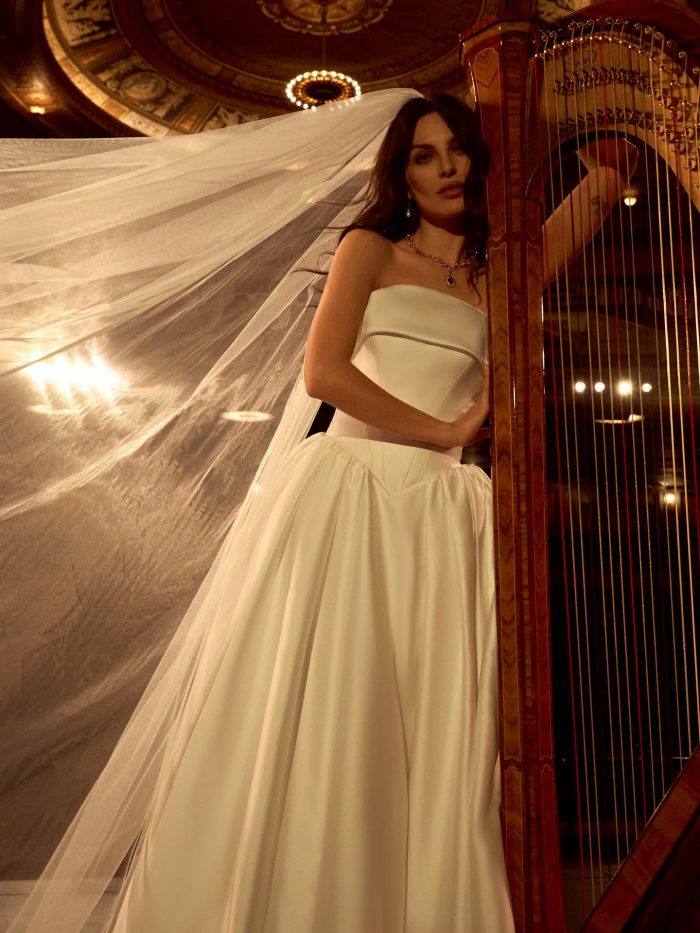 Close-up of a bride wearing the Rane ballgown wedding dress by Maggie Sottero, showcasing the strapless structured bodice and voluminous skirt, standing beside a harp in an ornate theater.
