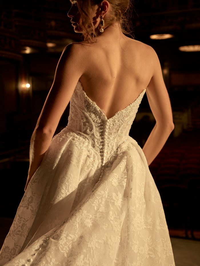 Back view of a bride wearing the Ryland ballgown wedding dress by Maggie Sottero, showcasing the beaded lace corset bodice with button-back detail and full lace skirt, in an ornate theater.