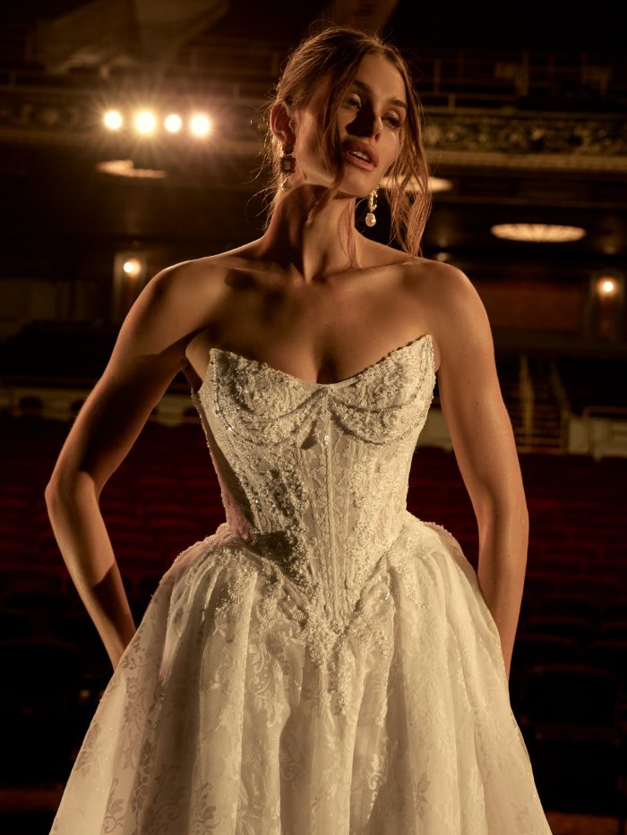 Close-up of a bride wearing the Ryland ballgown wedding dress by Maggie Sottero, featuring a strapless beaded lace bodice with sweetheart neckline, standing in an ornate theater.