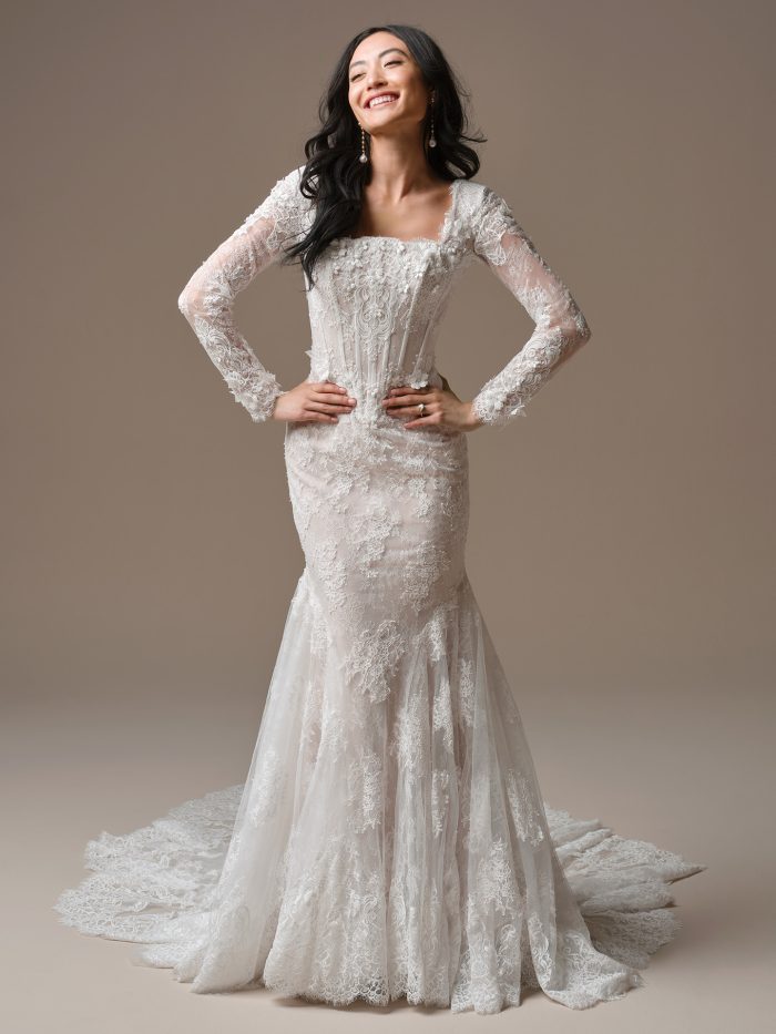 Takera Leigh wedding dress featuring Chantilly lace, pearl-accented 3D floral appliqués, a modest square neckline, long illusion lace sleeves, a fitted bodice with exposed boning and basque waistline, and a softly flared skirt with an extended scalloped illusion lace train. Model stands facing forward with hands on hips.