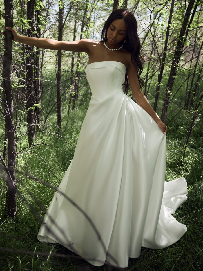 Bride wearing the Agatha A-line wedding dress by Rebecca Ingram, featuring a strapless structured bodice and full satin skirt, standing among trees in a wooded setting.