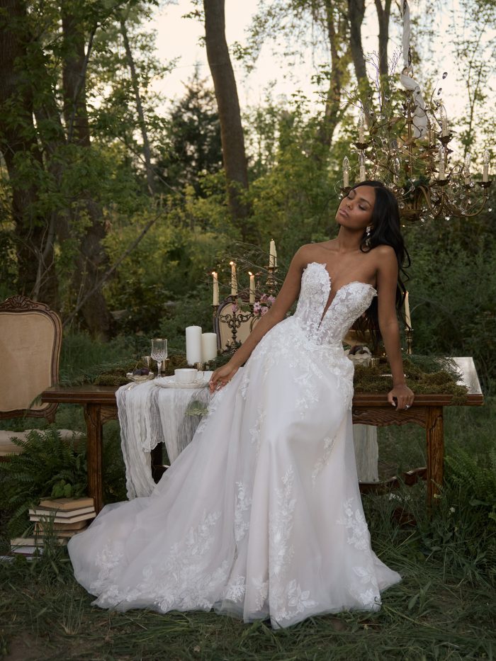 Bride wearing the Bernadine A-line wedding dress by Rebecca Ingram, showcasing the floral lace bodice and full tulle skirt with lace appliqués, seated at a candlelit table in a wooded outdoor setting.