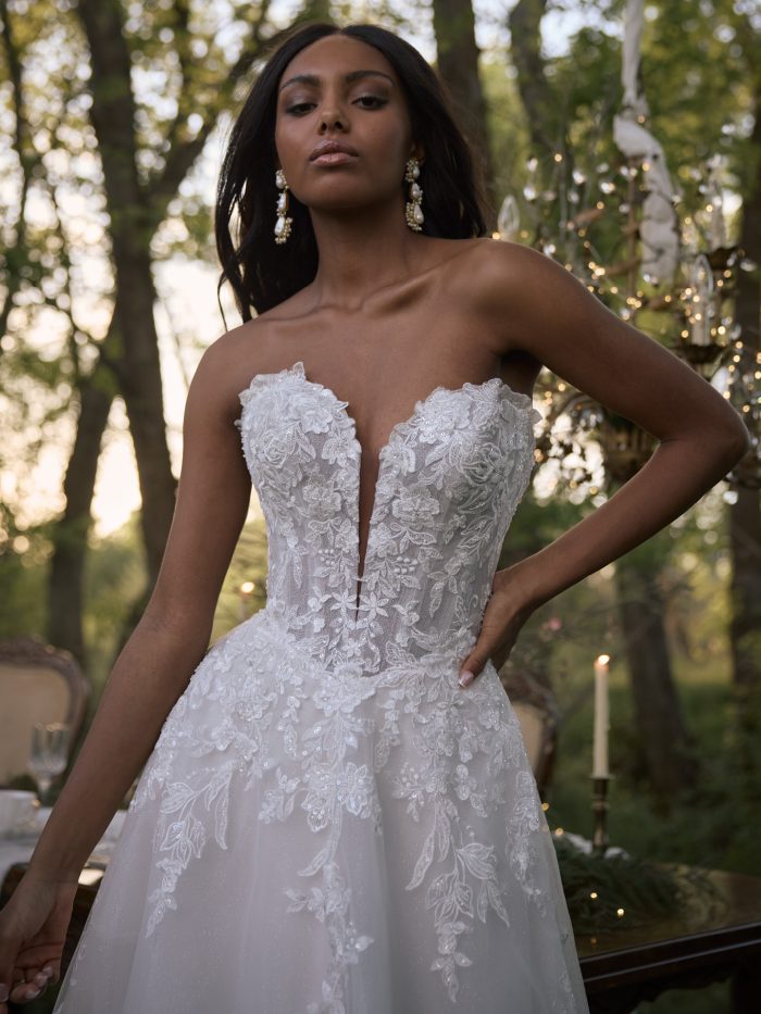 Close-up of a bride wearing the Bernadine A-line wedding dress by Rebecca Ingram, featuring a strapless sweetheart neckline with plunging floral lace bodice, in an outdoor evening setting with string lights.