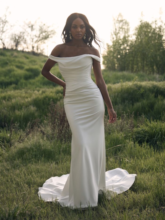 Bride wearing the Clementine sheath wedding dress by Rebecca Ingram, featuring an off-the-shoulder neckline, ruched bodice, and sweeping train, standing in a sunlit grassy field.