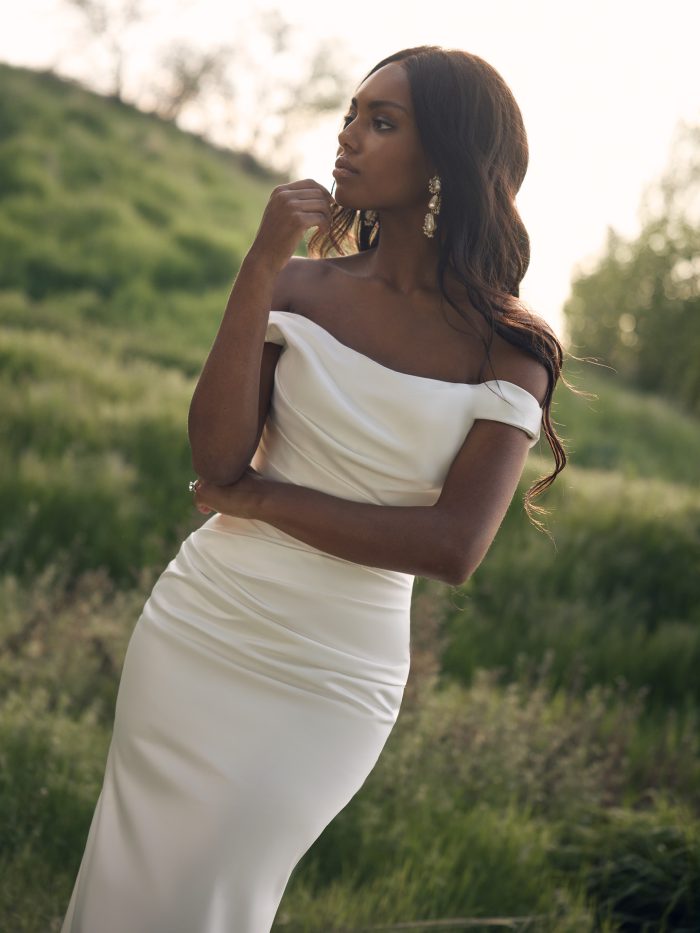 Close-up profile view of a bride in the Clementine sheath wedding dress by Rebecca Ingram, showcasing the asymmetrical off-the-shoulder neckline and fitted bodice detail.