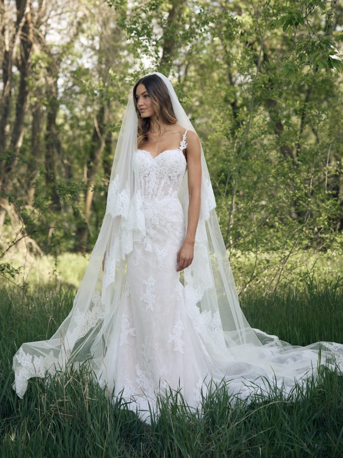 Bride wearing the Esmeralda Lane fit-and-flare wedding dress by Rebecca Ingram, showcasing the illusion lace bodice, fitted lace skirt, and cathedral veil, standing in a sunlit forest clearing.