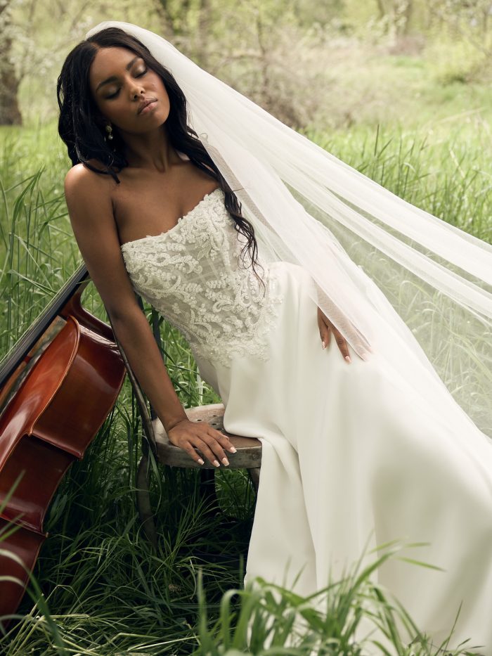 Bride wearing the Hazel A-line wedding dress by Rebecca Ingram, featuring a beaded lace strapless bodice and flowing veil, seated in a grassy field beside a cello.