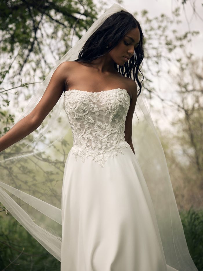 Close-up of a bride wearing the Hazel A-line wedding dress by Rebecca Ingram, showcasing the beaded lace bodice and full A-line skirt, with a flowing veil in an outdoor wooded setting.
