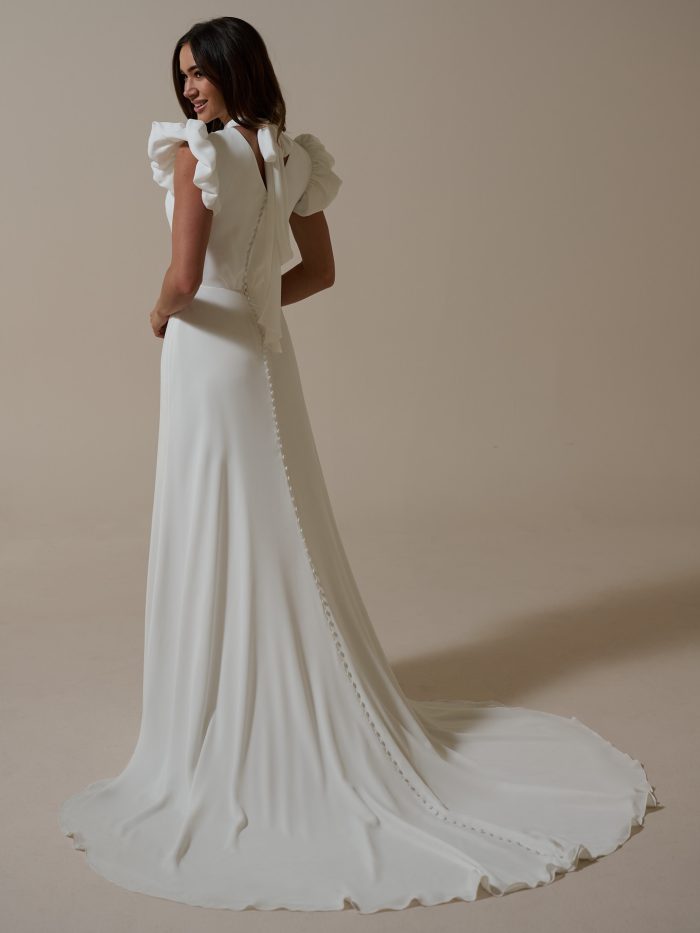 Back view of Payson by Rebecca Ingram, chiffon A-line wedding dress featuring ruffled puff sleeves, bow tie back, and row of covered buttons trailing to the train.