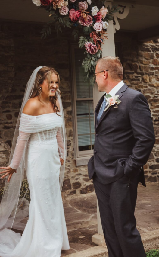 Bride in an off-the-shoulder pearl-embellished wedding dress with sheer long sleeves and veil, smiling at father in a dark suit under a floral arch at a rustic stone venue.