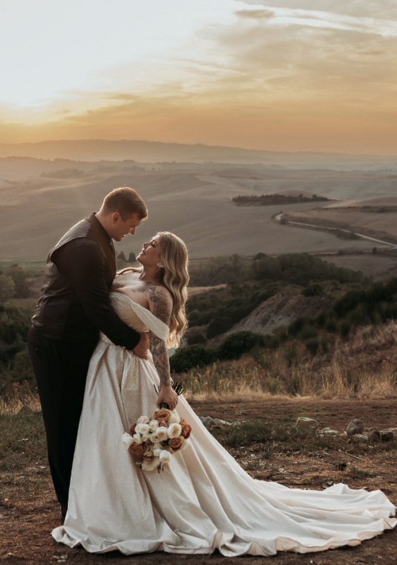 Bride in an off-the-shoulder satin ball gown with long train, holding a bouquet of neutral-toned flowers, sharing a romantic sunset moment with groom against rolling hillside views.