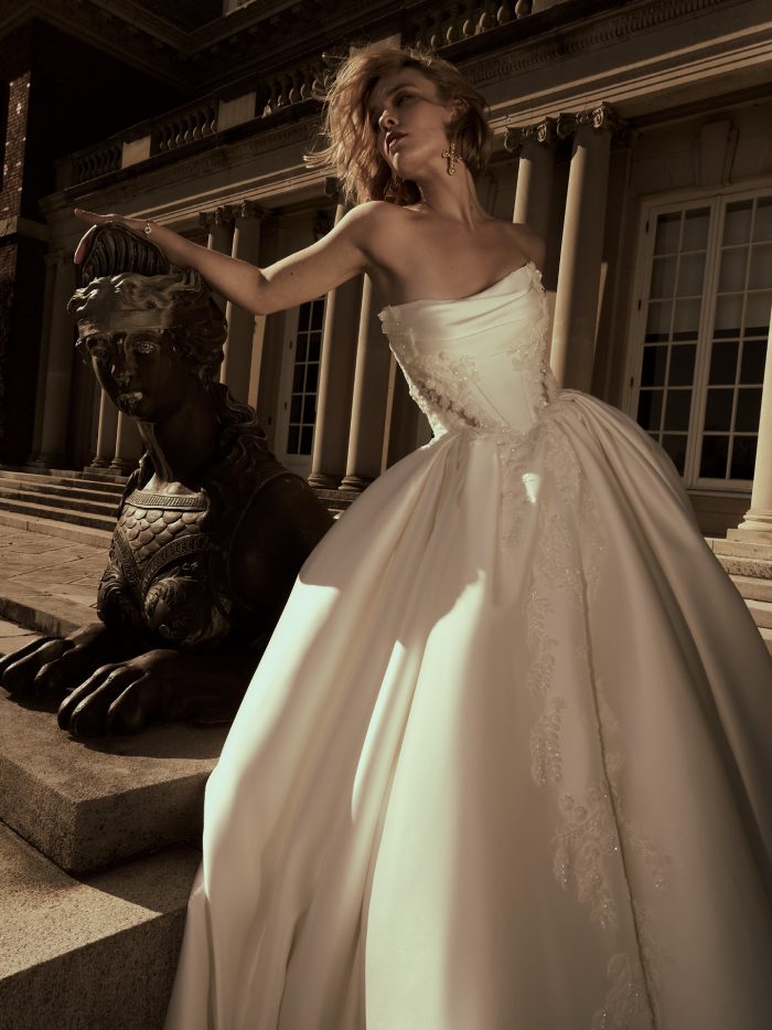Bride wearing the Burke Lane ballgown wedding dress by Sottero and Midgley, featuring a strapless draped bodice with lace appliqué detail and full satin skirt, posed beside a sphinx statue outside a grand building.
