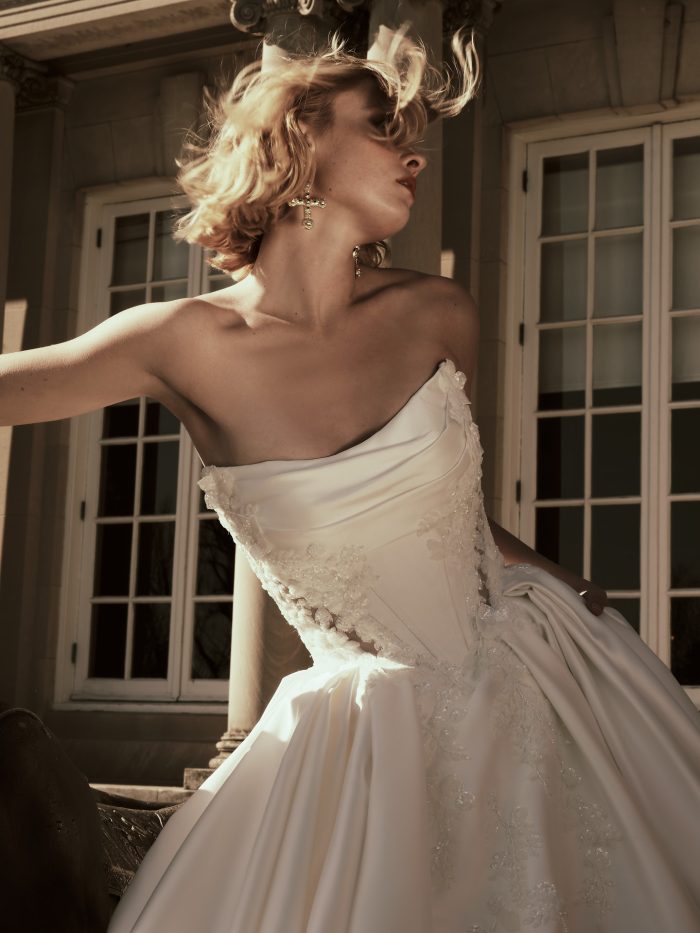 Close-up of a bride wearing the Burke Lane ballgown wedding dress by Sottero and Midgley, showcasing the draped strapless bodice with beaded lace appliqués, outside a grand columned building.